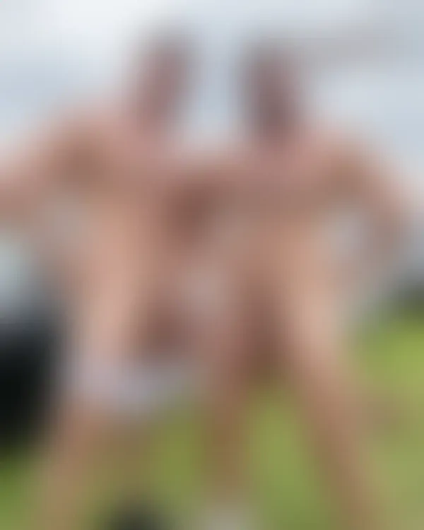 Two shirtless muscular men smiling confidently in a grassy mountain meadow under an open sky.