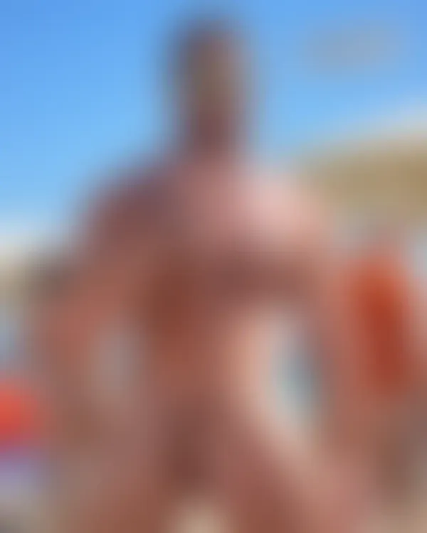 Muscular shirtless man on sunlit beach wearing mirrored sunglasses with hand near neck