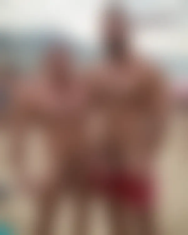 Two shirtless muscular men standing close on a sunny beach with arms around each other’s shoulders, smiling confidently 