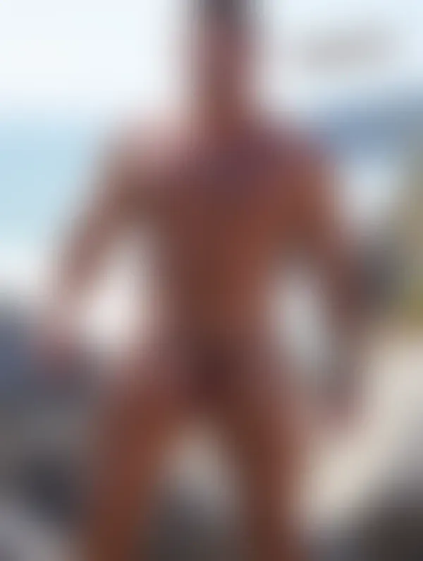 A muscular man with a silver cross necklace stands on coastal rocks under bright sunlight with the ocean roaring behind 