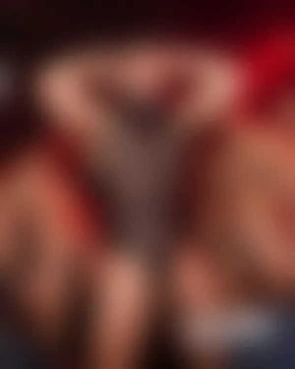 Muscular bearded man in sheer black mesh flexing confidently under saturated red club lights with tattooed companions.