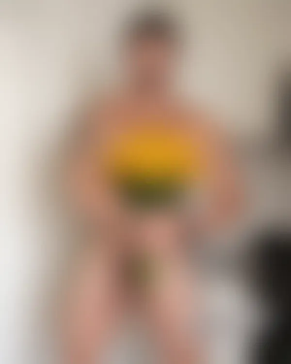 Shirtless muscular man holding a bouquet of yellow flowers against white kitchen cabinetry with an intense gaze and visi
