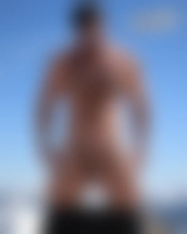 Shirtless tattooed man with sunglasses standing on a snowy mountain peak under a clear blue sky
