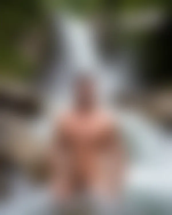 Muscular tattooed man waist-deep in rushing mountain stream with cascading waterfall and lush green forest behind him.