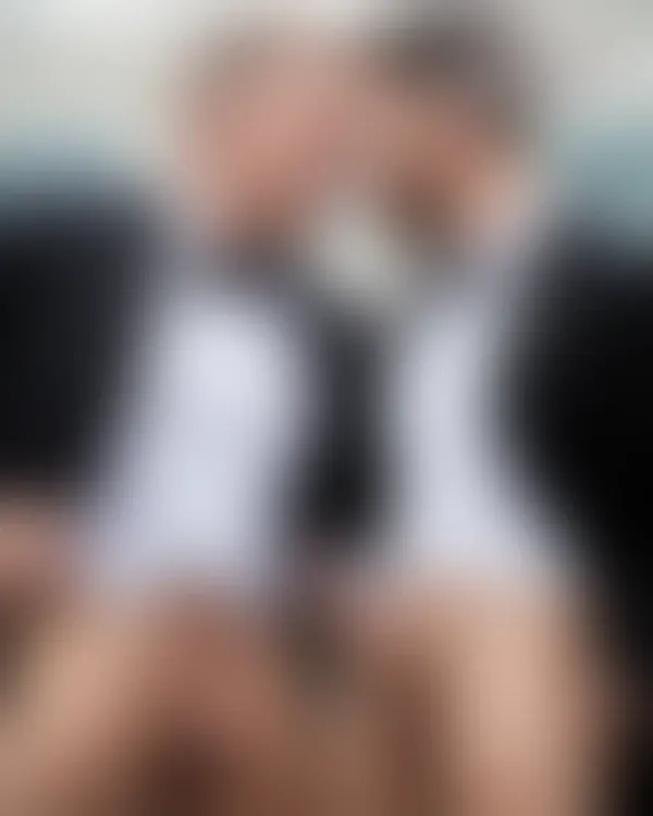 Two men in tuxedos leaning forehead to forehead with eyes closed; suits slightly undone at the waist against a blurred u