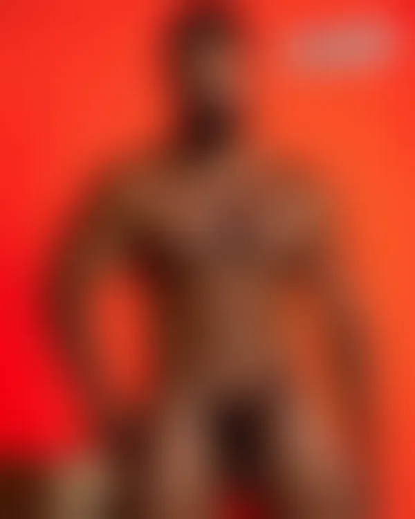 Muscular bearded man shirtless under red lighting with intense direct gaze and defined torso muscles