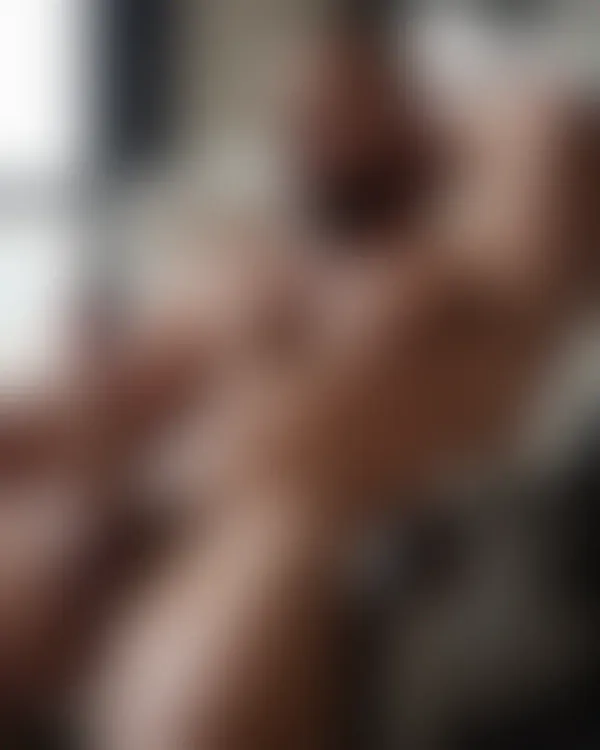 Muscular bearded man shirtless lounging in a cream armchair under soft natural light with an intense gaze