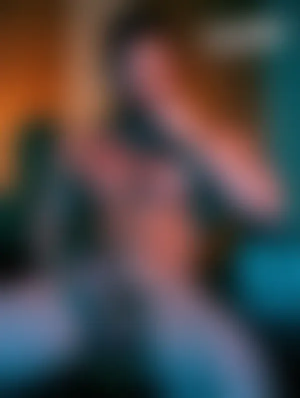 A muscular bearded man in a dim teal-lit room, shirtless, gazing directly at the camera with raw confidence