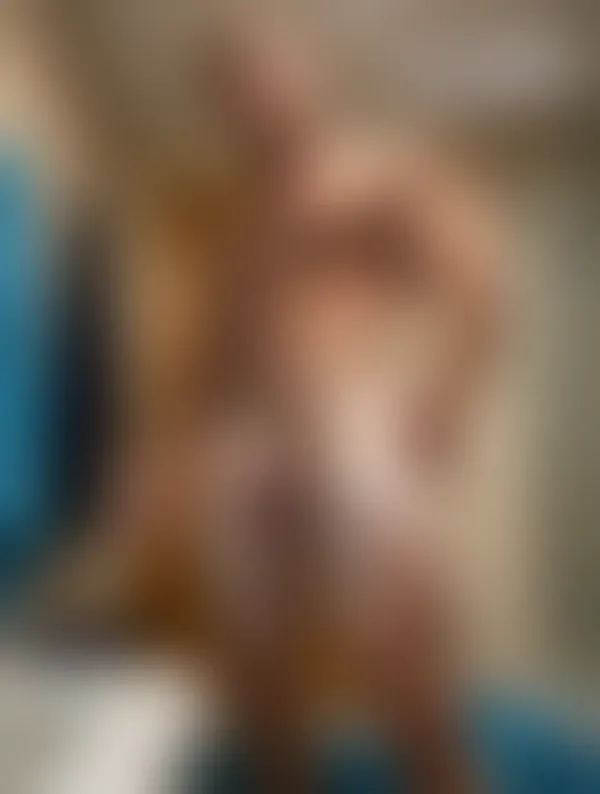 A muscular man covered in white soap suds standing nude in a tiled bathroom and gazing directly at the camera.