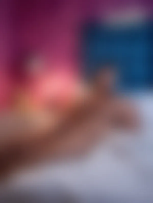A muscular man reclining nude on a bed against a blue plaid headboard, hand near his erect penis, pink toiletry…