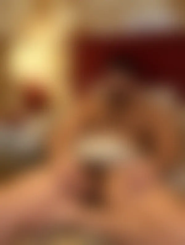 Muscular shirtless man reclining on a bed with red headboard, hands near exposed genitalia in an intimate indoor setting
