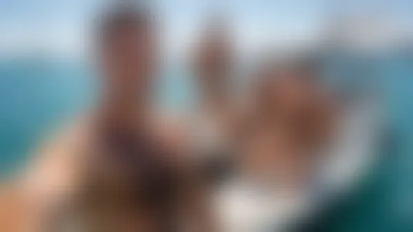 Four shirtless muscular men posing on a sunny sailboat with turquoise water and distant boats behind them.